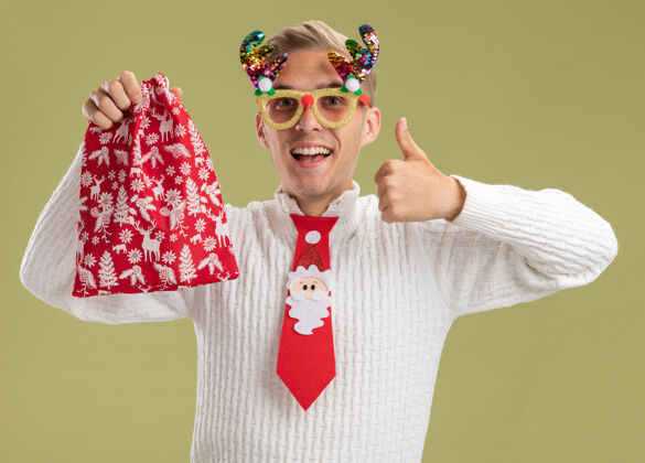 向上令人印象深刻的年轻帅哥戴着圣诞新奇眼镜 打着圣诞老人的领带 拿着圣诞袋 看着相机 在橄榄绿的背景上孤立地竖起大拇指显示圣诞老人年轻