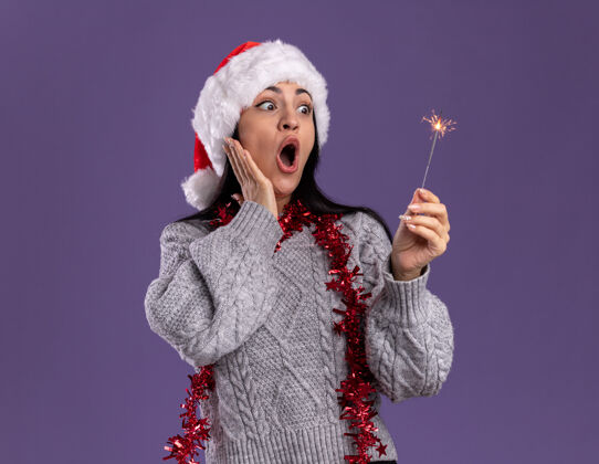 圣诞快乐令人印象深刻的年轻白种女孩戴着圣诞帽 脖子上戴着金箔花环 手放在脸上 手放在紫色背景上 看着节日的火花花环复制空间
