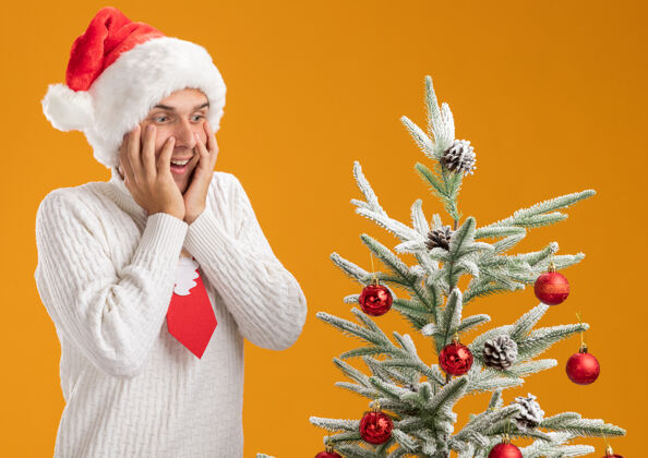 站起来令人印象深刻的年轻帅哥戴着圣诞帽 打着圣诞老人的领带 站在装饰好的圣诞树旁 手放在脸上 在橙色的背景下孤立地往下看年轻人圣诞老人圣诞老人
