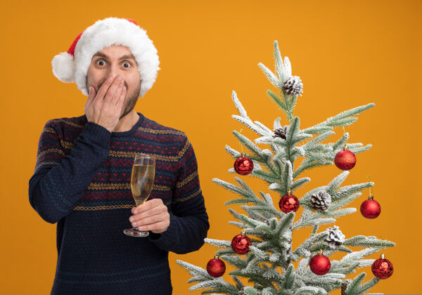 新年一个戴着圣诞帽的白人年轻人站在装饰好的圣诞树旁 手里拿着一杯香槟 手放在嘴上 看着橙色背景下的摄像机关心圣诞香槟