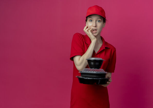 印象令人印象深刻的年轻漂亮的送货女孩穿着红色制服 戴着帽子拿着食品容器 咬着她的手指在深红色的背景与复制空间隔离帽子持有红色
