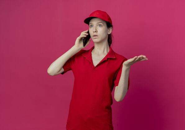 背景令人印象深刻的年轻漂亮的送货女孩穿着红色制服 戴着帽子打电话 看起来笔直 显示空的手在深红色的背景与复制空间隔离制服显示深红色