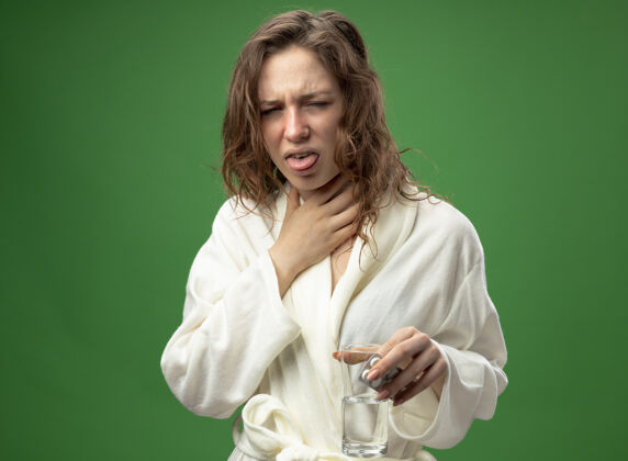 穿咳嗽的年轻病女孩穿着白色长袍 手里拿着一杯水 掐住了隔离在绿色上的喉咙袍年轻抓