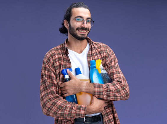 年轻微笑的年轻帅气的清洁工穿着t恤拿着清洁工具隔离在蓝色的墙上抱着工具小伙子