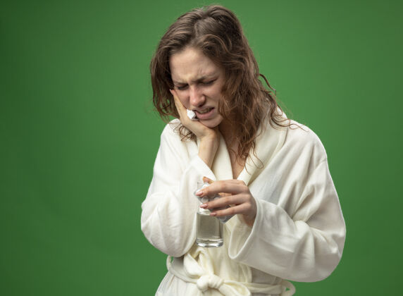 病疼痛的年轻病女孩穿着白色长袍拿着一杯水把手放在脸颊上隔离在绿色颊绿穿