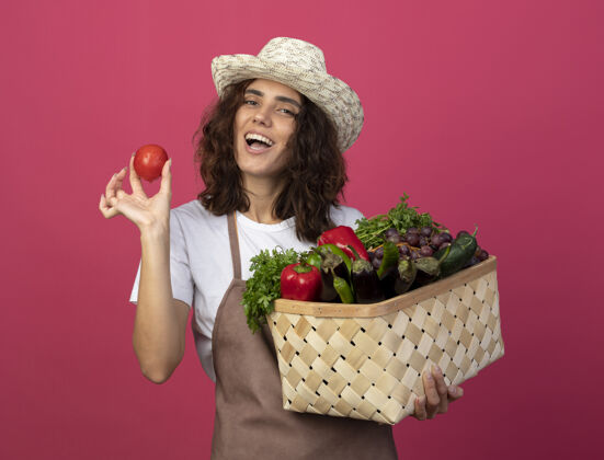 年轻快乐的年轻女园丁穿着制服 戴着园艺帽 手里拿着蔬菜篮子 篮子里的西红柿被隔离在粉色上篮子帽子欢乐