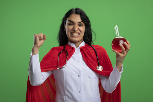 年轻兴奋的年轻超级英雄女孩穿着医用长袍 手持听诊器 拿着装满红色液体的化学玻璃瓶 在绿色墙上显示“是”的手势听诊器填补举行
