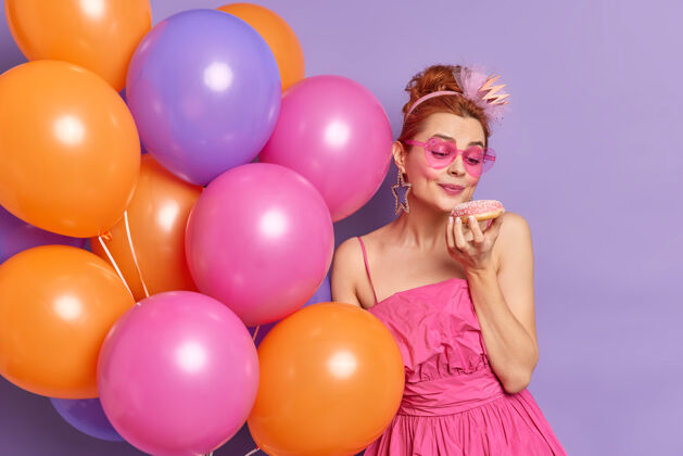 太阳镜人们节日和庆祝的概念可爱的红发年轻女子看着开胃的甜甜圈举行了紫色背景上孤立的彩色气球一串一美味女性