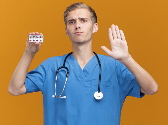 男性严格的年轻男性医生穿着医生制服 手持听诊器 在橙色墙上显示停止手势医生戴着年轻