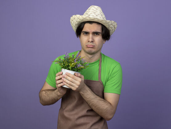 帽子悲伤的年轻男园丁穿着制服戴着园艺帽捧着花盆里的花孤立在紫色的花坛上花悲伤年轻