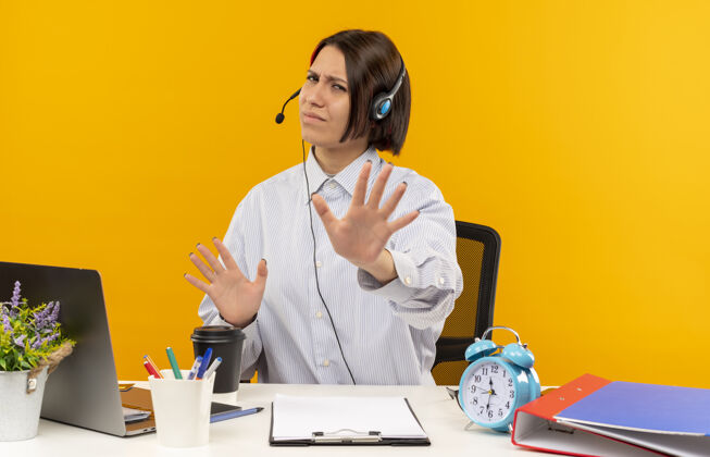 耳机未经许可的年轻呼叫中心女孩戴着耳机坐在办公桌旁 工作工具没有做任何手势 孤立在橙色背景上女孩手势穿着