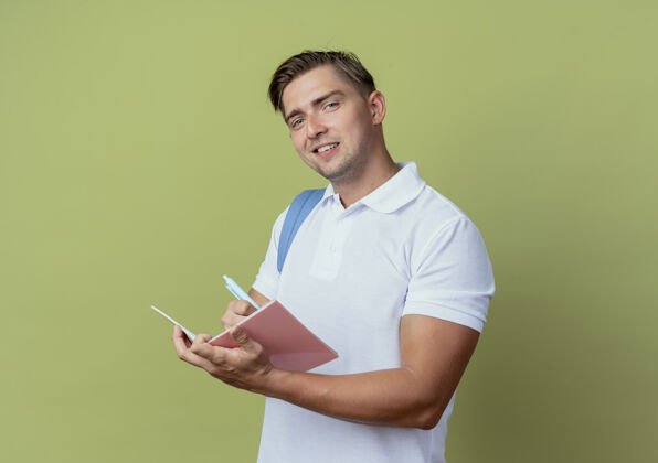 橄榄高兴的年轻英俊的男学生戴着背袋写东西在笔记本上孤立的橄榄绿背景穿什么书包写字