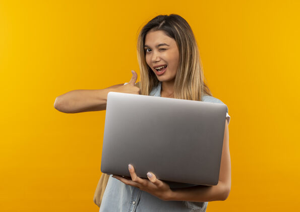 背部快乐的年轻漂亮的女学生戴着背袋拿着笔记本电脑 拇指向上 眨眼孤立的橙色眨眼包橙色