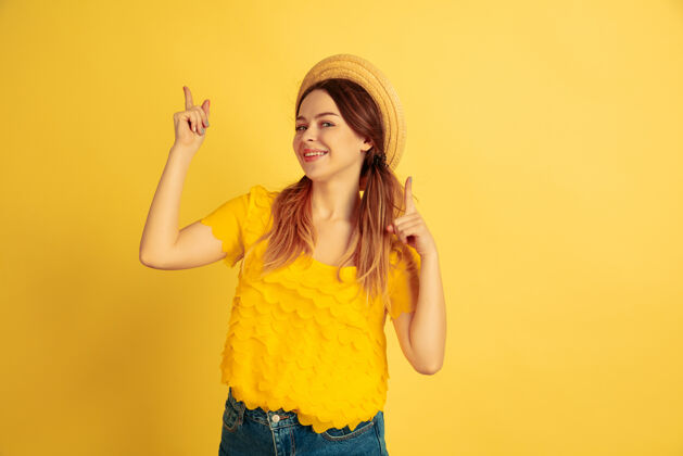 夏季指着 微笑黄色工作室背景上的白人女性肖像戴着帽子的漂亮女模特人类情感的概念 面部表情 销售 广告夏季 旅游 度假年轻人情感