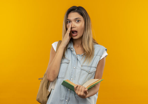 背印象深刻的年轻漂亮的学生女孩穿着背袋拿着打开的书把手放在嘴边低语在前面孤立的橙色年轻近开
