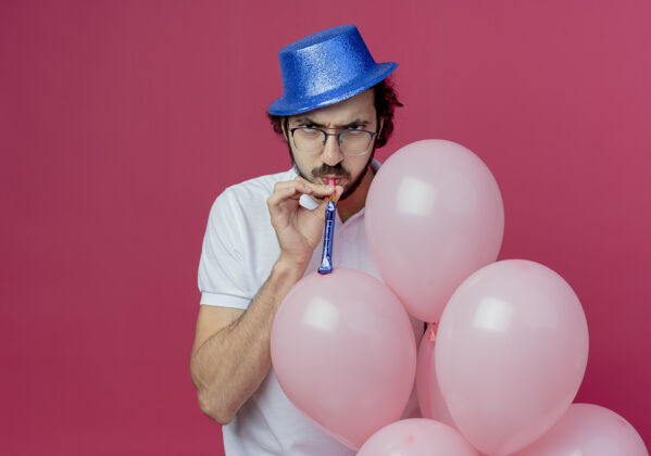 气球一个戴着眼镜 戴着蓝色帽子 手持气球 吹着哨子的严厉帅哥孤立在粉色背景上手持穿严格