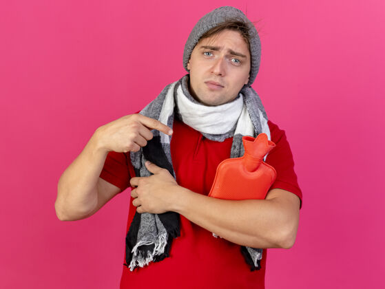 人年轻英俊的金发病男子戴着冬天的帽子和围巾拿着和指着热水瓶看着前面孤立的粉红色墙壁上的复制空间衣服人穿着