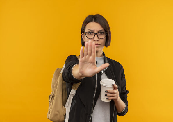 年轻未出租的年轻学生女孩戴着眼镜 背着提包拿着咖啡杯 在橙色背景下的相机前做手势 并留有复印空间眼镜杯子停止