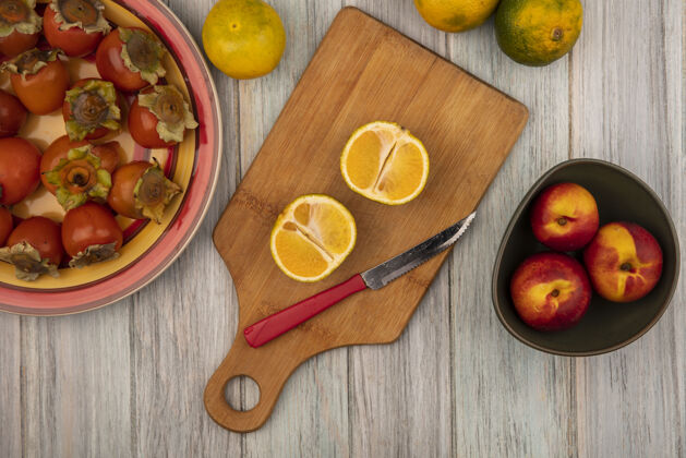 刀木制厨房板上的有机新鲜橘子的俯视图 刀子上放着桃子 盘子上放着柿子 背景是灰色木制的桃柑橘健康