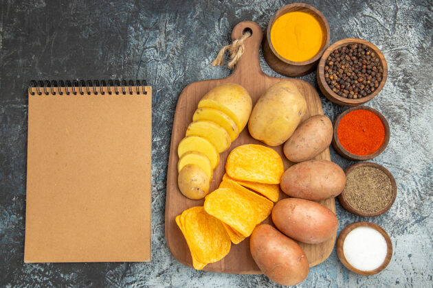 香料上图：脆片和生土豆放在木制砧板上不同的香料和灰色桌子上的笔记本蔬菜膳食纸板