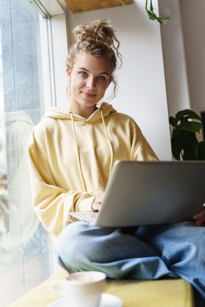 桌子坐在窗户上 把笔记本电脑放在膝盖上 美丽微笑的女人用笔记本电脑的垂直镜头金发聊天电脑