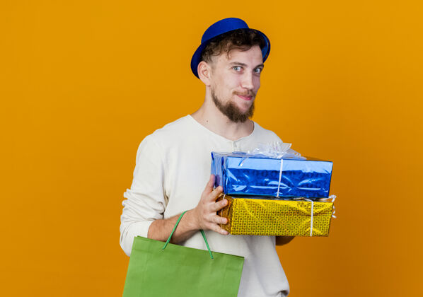 橙色高兴的年轻英俊的斯拉夫党人戴着党的帽子拿着礼品盒和纸袋看着相机孤立的橙色背景与复制空间包礼物斯拉夫人