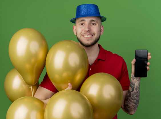帽子面带微笑的年轻帅哥斯拉夫党的家伙戴着党的帽子站在气球后面展示手机看着相机隔离在绿色背景表演手机站立