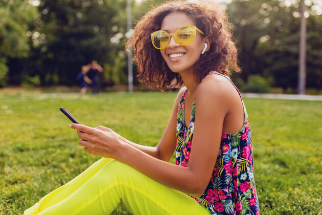 女孩年轻时尚微笑的黑人女子用智能手机听音乐无线耳机在公园里玩得开心 夏日时尚多彩的风格 坐在草地上 戴着黄色太阳镜人休闲电话