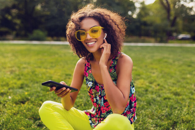 时尚年轻时尚微笑的黑人女子用智能手机听音乐无线耳机在公园里玩得开心 夏日时尚多彩的风格 坐在草地上 戴着黄色太阳镜年轻耳机眼镜