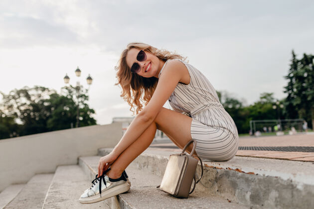 时尚年轻漂亮时尚的金发女郎坐在城市街道上 穿着夏日时尚的连衣裙 戴着太阳镜 钱包 银色运动鞋优雅鞋子腿