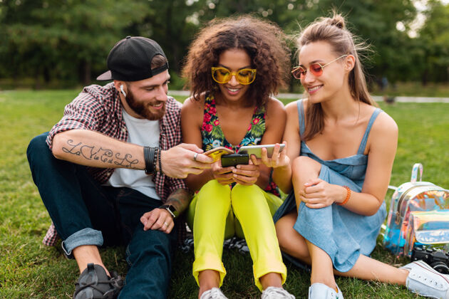 时尚快乐的年轻朋友们坐在公园里用智能手机微笑 男人和女人一起电话友谊