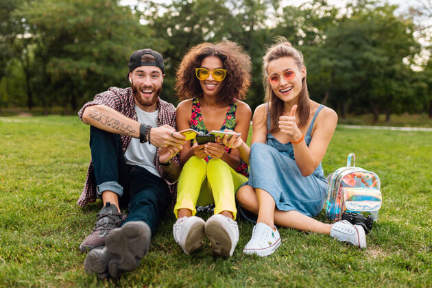 兴奋快乐年轻的朋友们坐在公园里用智能手机微笑 一起享受快乐多彩的夏日风格 通讯无线连接设备 在相机里看起来很积极时尚手机多种族