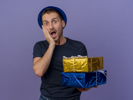 蓝色一个戴着蓝色帽子的白人帅哥惊讶地把手放在脸上 手里拿着一个紫色背景的礼品盒 上面留有复印空间脸紫色持有