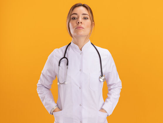 自信自信的年轻女医生穿着医用长袍 听诊器把手放在隔离在黄色墙上的口袋里年轻穿着医生