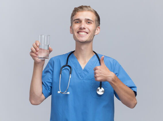 年轻微笑着的年轻男医生穿着医生制服 手持听诊器 举着一杯水 在白色的墙上孤立地竖起大拇指微笑医生男性