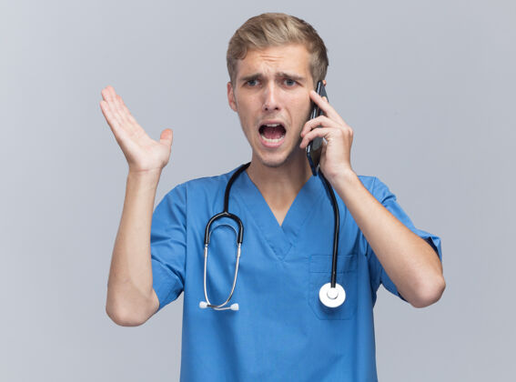 电话愤怒的年轻男医生穿着医生制服 手持听诊器在电话里讲话 手被隔离在白墙上制服愤怒传播