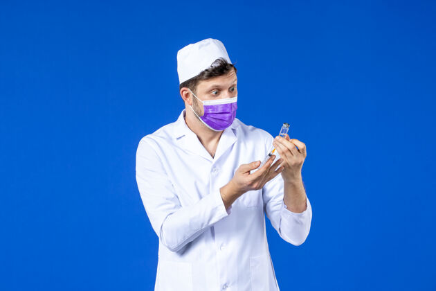 大流行前视图中的男医生穿着医疗服和口罩持有疫苗和注射在蓝色注射西装外套