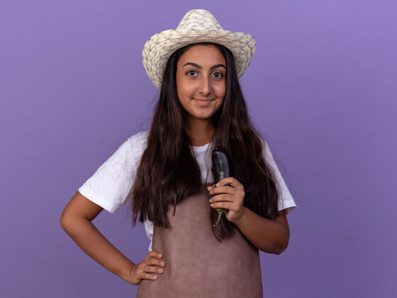 立场穿着围裙 戴着夏帽 抱着茄子的年轻园丁女孩站在紫色的墙上 快乐而积极地微笑着积极的茄子花园
