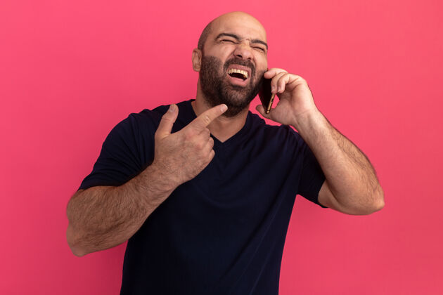 胡子一个留着大胡子的穿海军t恤的男人站在粉色的墙上对着手机大喊着“受挫”站着沮丧电话