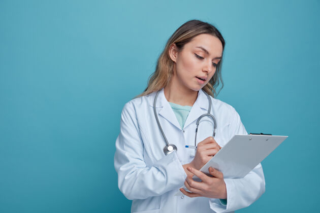 剪贴板印象深刻的年轻女医生穿着医用长袍和听诊器围着脖子用笔在剪贴板上写字蓝色女性脖子