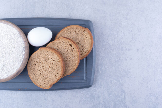农产品一小碗面粉 一个鸡蛋和三片面包放在大理石表面的海军蓝木板上酵母烘焙食品面团