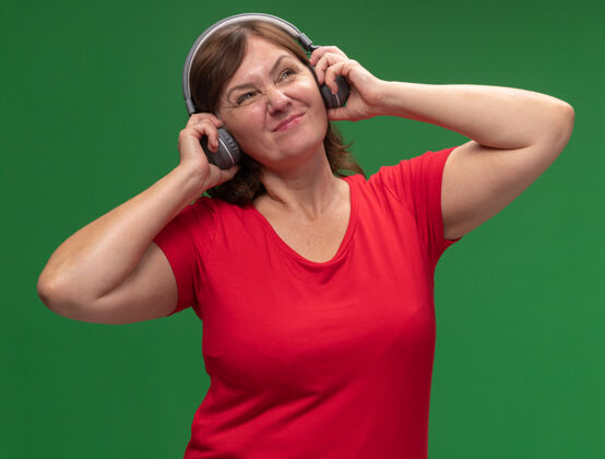 中年穿着红色t恤 戴着耳机听音乐的中年妇女站在绿墙边不高兴不高兴音乐耳机