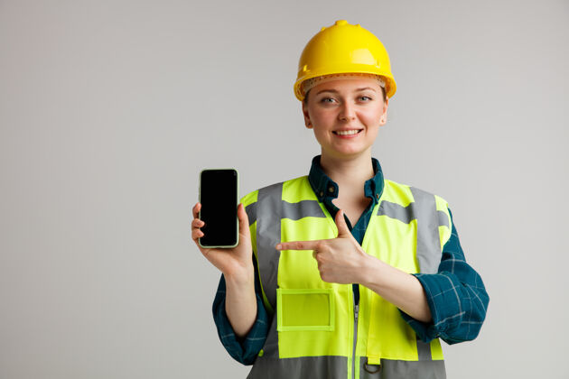 安全面带微笑的年轻女建筑工人戴着安全帽和安全背心手持并指着手机手机电话手持