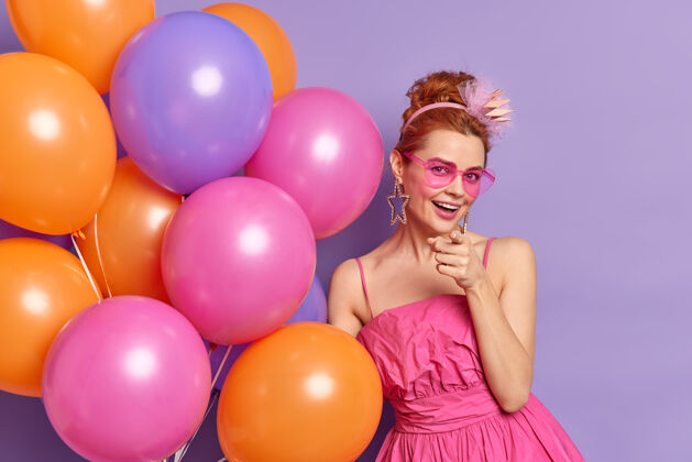 积极积极的红发女人在你的派对和庆典邀请上表示 戴着时髦的太阳镜 穿着盛装举着一堆充气气球庆祝周年情绪女士太阳镜