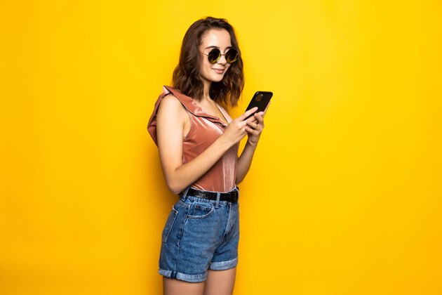 短信一个戴着夏天帽子和太阳镜的惊讶的年轻女子的肖像 用手机隔离在黄色的墙上手机女性电话