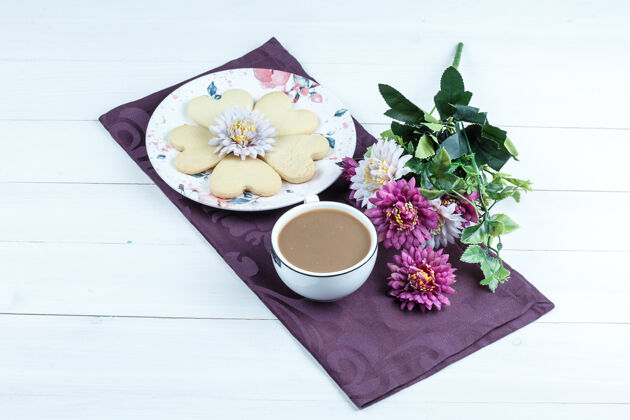 薯条心形饼干 一杯咖啡放在一张紫色垫子上 白色木板背景上有花 高角度观看花形状木板
