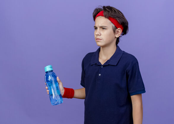 奶瓶皱眉的年轻帅气的运动男孩戴着头带和护腕 戴着牙套 拿着水瓶 看起来笔直地孤立在紫色的墙上 留着复制空间穿帅气运动