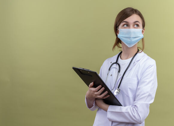 面具体贴的年轻女医生穿着医用长袍 戴着口罩 手持听诊器 拿着剪贴板 看着隔离的绿墙上有复印空间的右侧思想医生年轻人