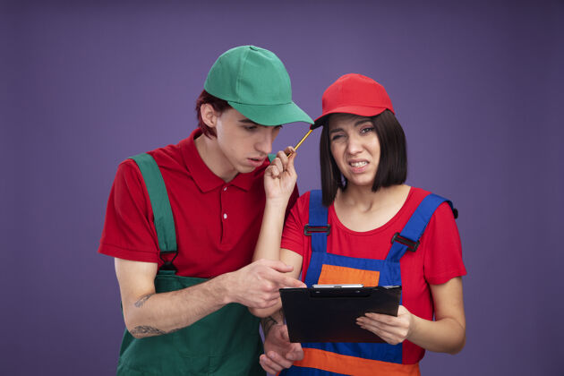 家伙一对穿着建筑工人制服 戴着帽子的年轻夫妇困惑地拿着铅笔和剪贴板的女孩用铅笔摸着头专心地看着并指着剪贴板制服夫妇帽子