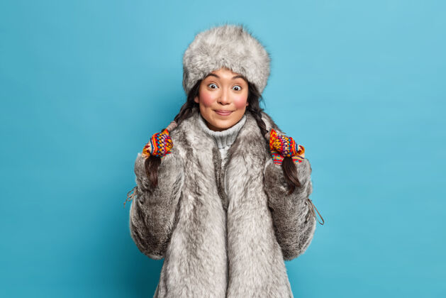 室内一个年轻的亚洲女人手上扎着辫子 穿着外套 隔着蓝色的墙爱斯基摩女人戴着帽子 穿着外套 住在北极地区欢呼辫子毛衣